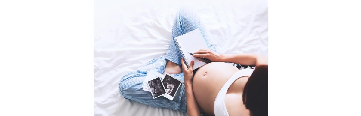 Mulher grávida sentada na cama usando top escrevendo em caderno e olhando fotos
