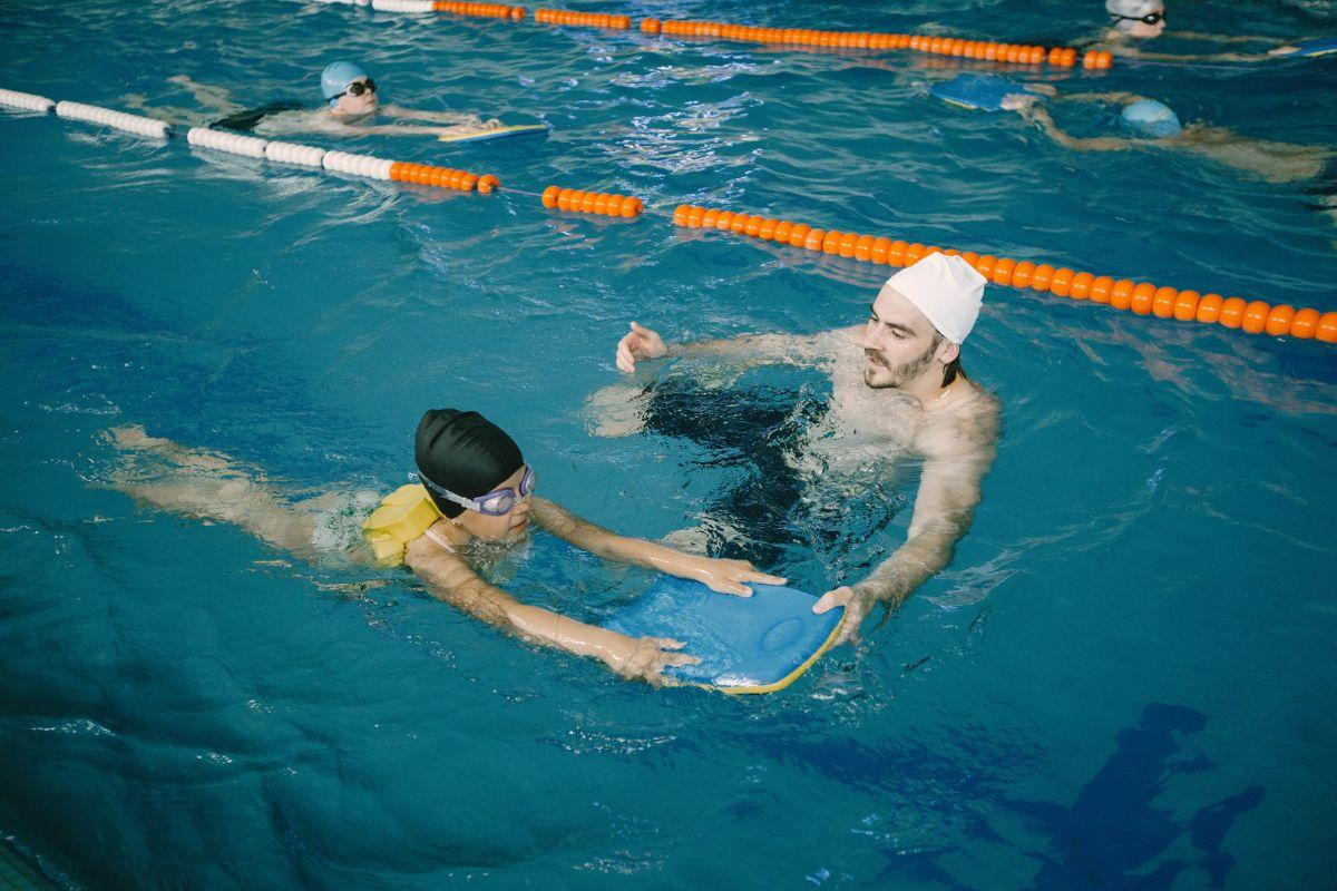 Homem de touca de natação segurando prancha com criança em piscina, em aula de natação